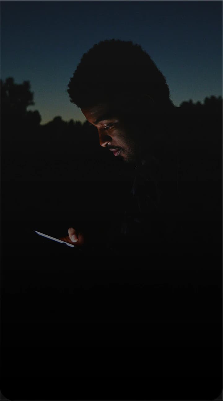 A man outdoors at dusk, his face lit by the phone in his hand