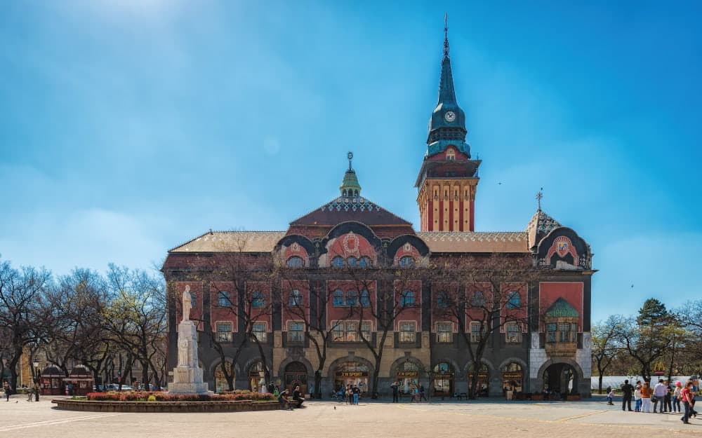 Stone statue standing in the middle of Liberty Square with the red-and-cream Art Nouveau City Hall building and its tall clock tower rising behind it under a clear blue sky