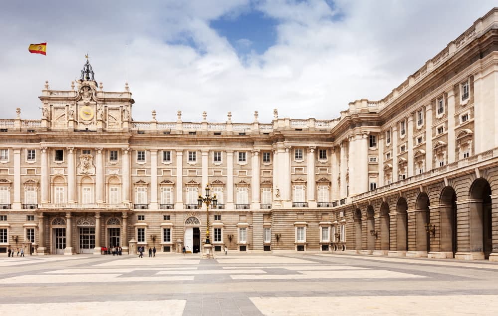 Royal Palace of Madrid exterior facade with Spanish flag, beige limestone neoclassical architecture, official ceremonial residence with ornate details in Spain's historic city center