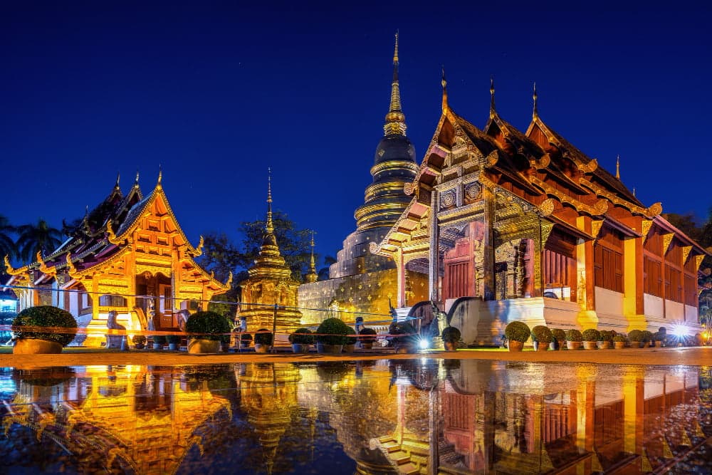 Night view of Wat Phra Singh temple in Chiang Mai, Thailand, with illuminated golden pagodas and traditional Lanna architecture reflecting in water.