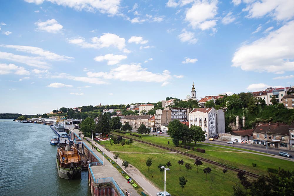 View along Belgrade’s Sava River quay showing docked boats, a paved promenade, grassy parkland with a disused railway track, and hillside buildings topped by the Church of Saint Michael the Archangel’s ornate bell tower under a partly cloudy sky.