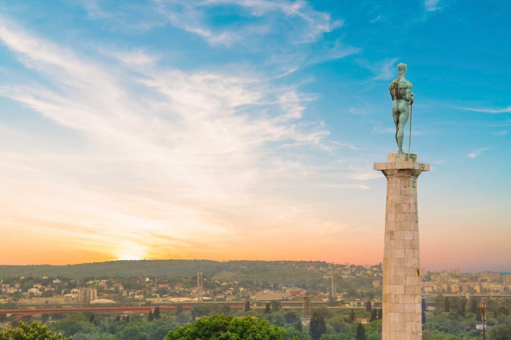 Bronze statue of a standing male figure holding a sword and falcon atop a tall stone column, overlooking the city of Belgrade and the river below at sunset.
