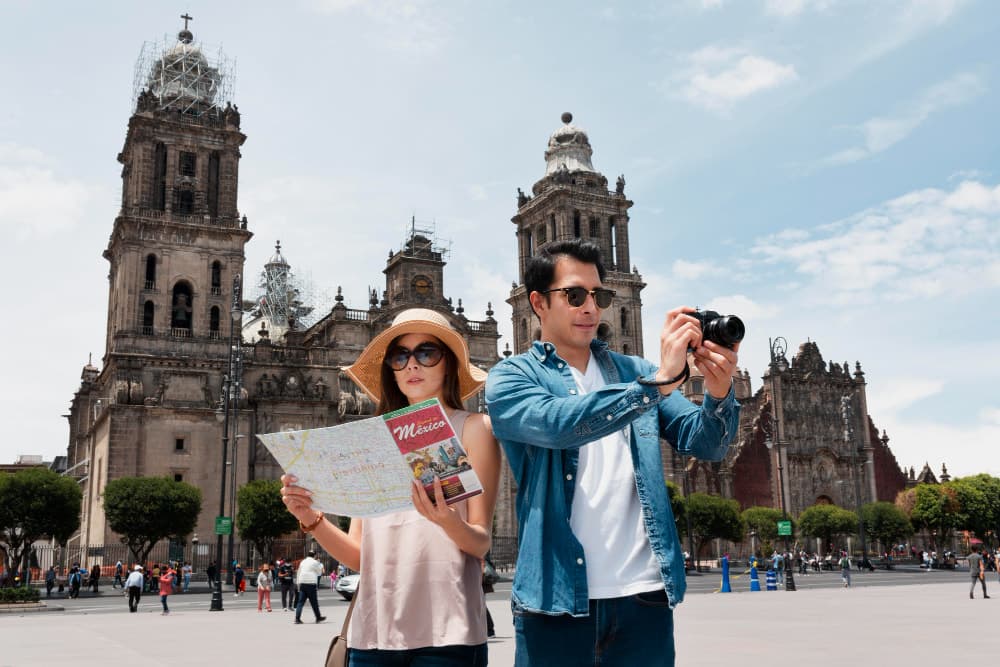 Tourists exploring the Zócalo plaza in Mexico City with the historic Metropolitan Cathedral in the background, one reading a map and the other taking photographs.