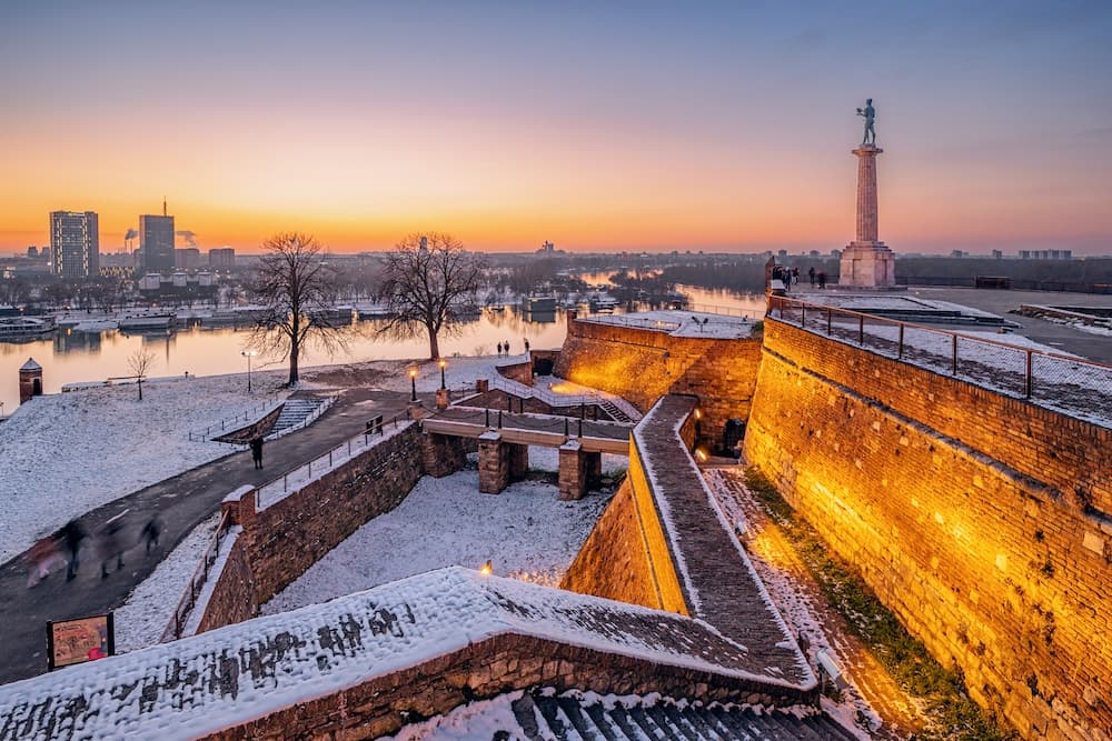 Snow-dusted brick ramparts and stairways of Belgrade Fortress leading up to the Pobednik monument atop its column, with the Sava River and city skyline glowing in the winter sunrise.