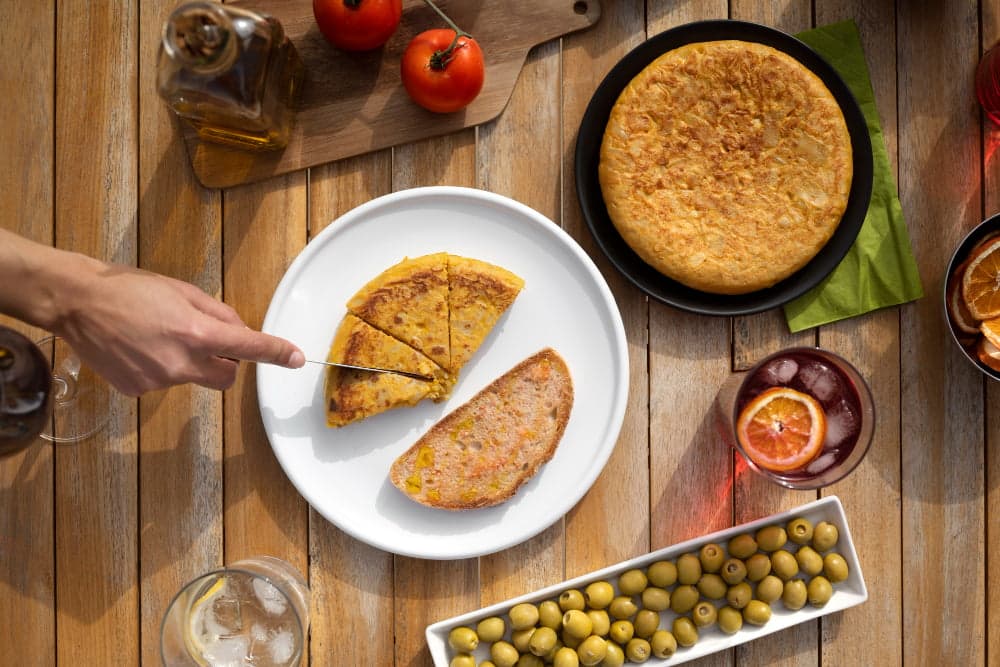 Traditional Spanish tortilla de patatas on white plate with tapas spread including green olives, Spanish omelette, olive oil, tomatoes, and sangria on wooden table, authentic Mediterranean cuisine