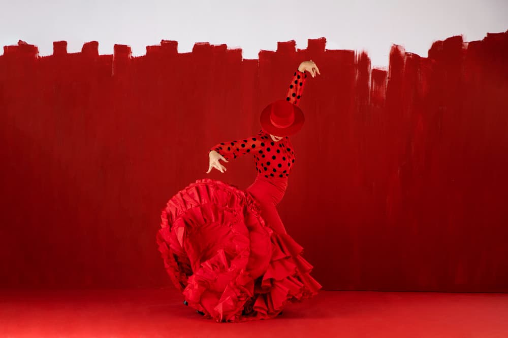 Spanish flamenco dancer in traditional red traje de flamenca dress with polka dots and ruffles performing dramatic pose against vibrant red background, authentic Andalusian cultural performance art