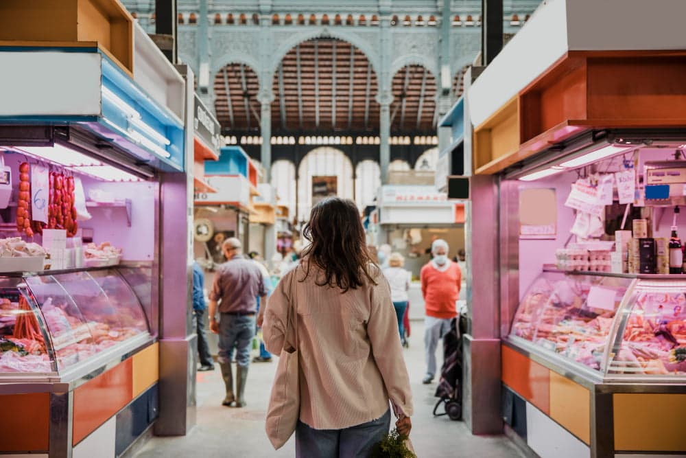 Historic Spanish food market interior in Madrid with person walking between meat stalls, colorful vendor displays, traditional Moorish architectural arches, and bustling shopping atmosphere in Mercado central