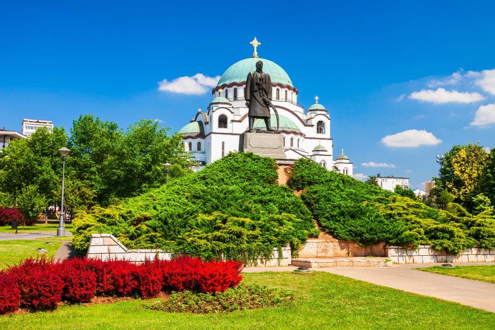 Statue of Grand Prince Stefan Nemanja standing atop a landscaped mound in front of the white-marble, green-domed Temple of Saint Sava on Vračar plateau in Belgrade under a clear blue sky.