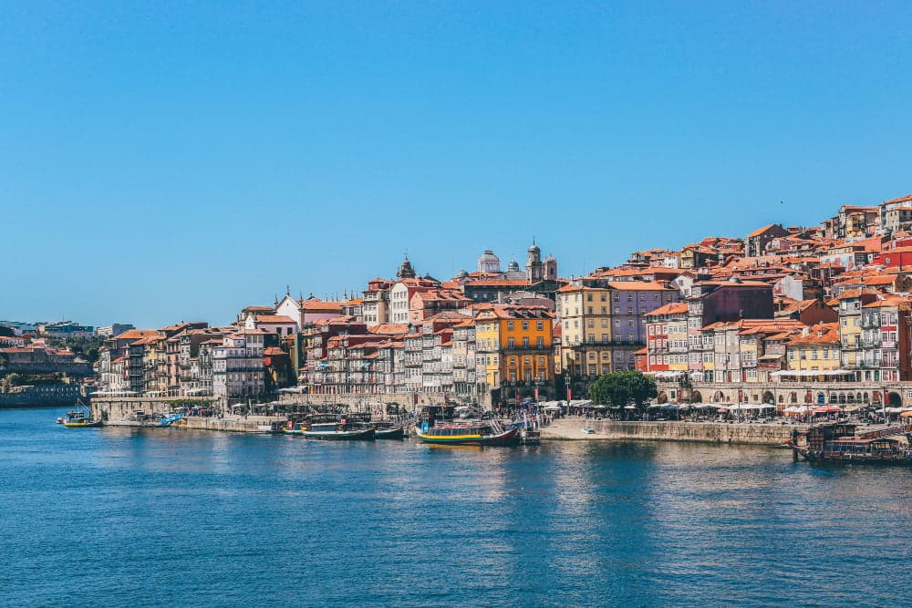 Colorful historic buildings along the Douro River waterfront in Porto, Portugal, with traditional boats and a clear blue sky.
