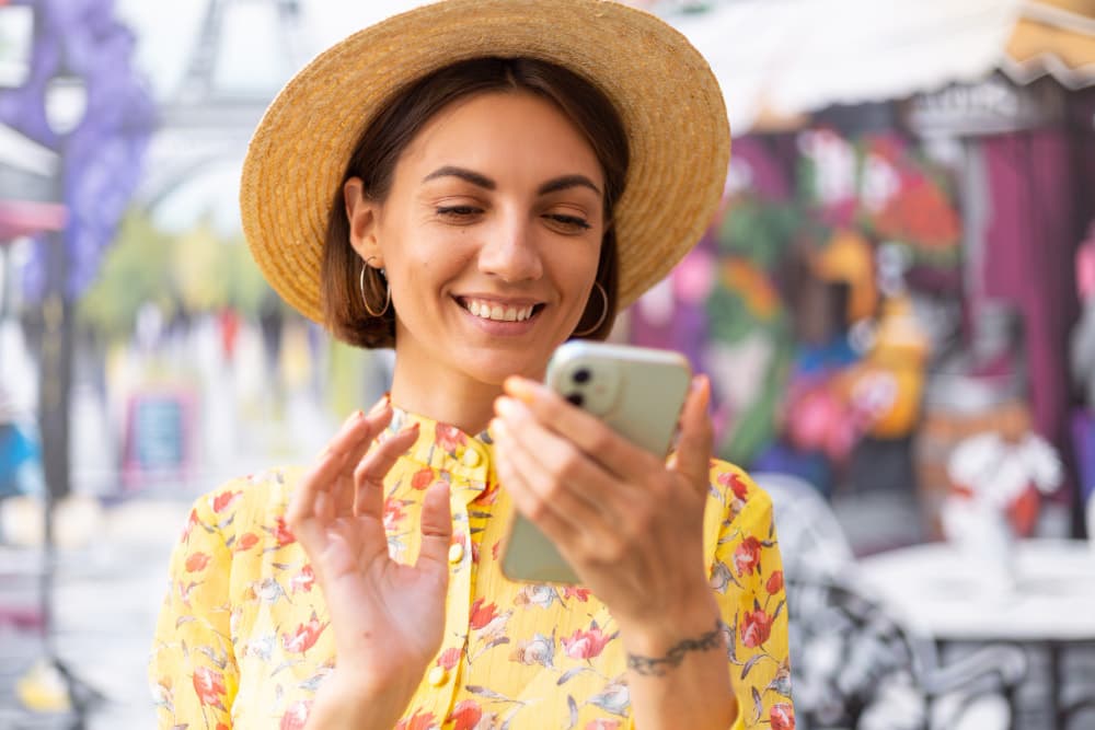 Woman wearing a straw hat and floral yellow dress using a smartphone outdoors in a vibrant, colorful street cafe setting.