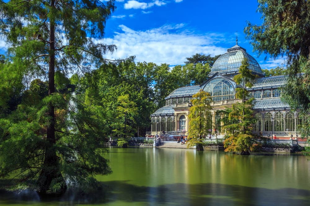 Crystal Palace in Madrid's Retiro Park, historic glass and metal greenhouse structure reflecting in artificial lake, contemporary art exhibition venue surrounded by lush green trees under blue sky