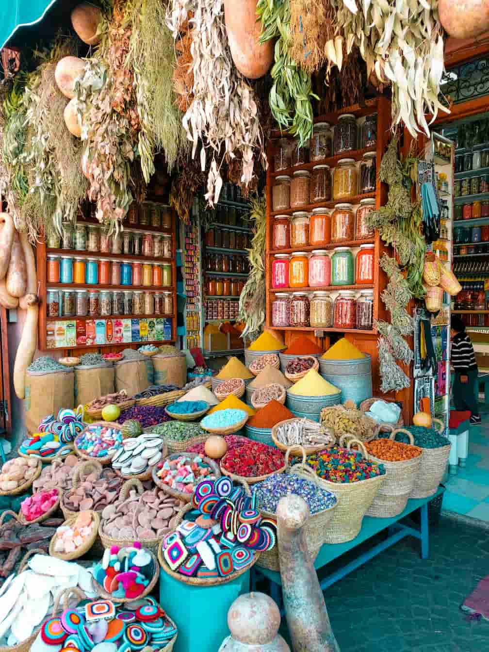 A vibrant Moroccan spice market with colorful spices, herbs, and dried goods displayed in baskets and jars, surrounded by hanging dried plants.