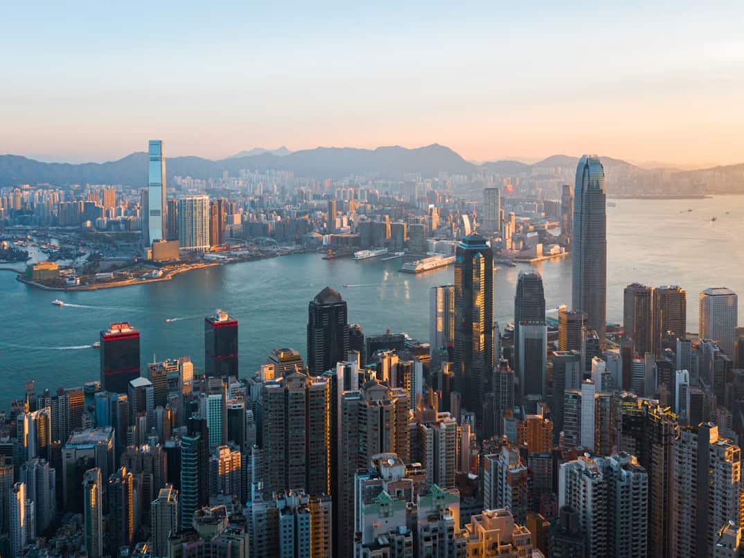 Aerial view of a bustling cityscape with numerous skyscrapers and buildings along a waterfront, under a clear blue sky at sunset.