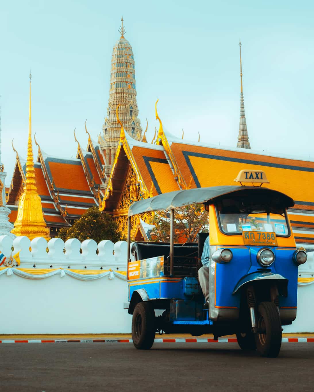 A traditional tuk-tuk taxi is parked in front of an ornate temple with golden spires under a clear blue sky.