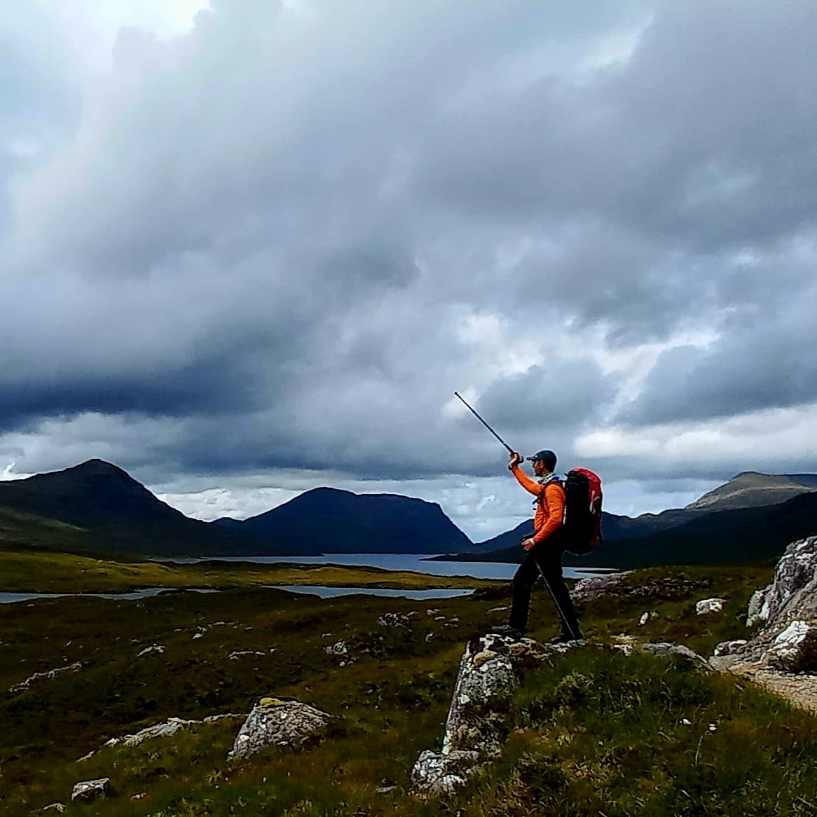 Hiker in an orange jacket holds a walking stick, standing on a rock with mountains and a cloudy sky in the background.