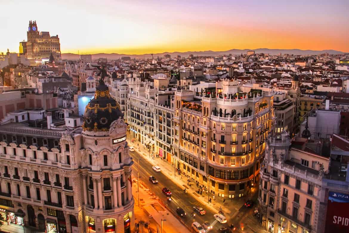 Aerial view of a vibrant cityscape at sunset, with illuminated historic buildings lining a bustling street and a mountainous horizon in the distance.