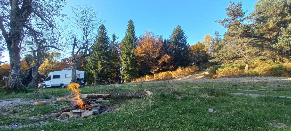 A camper van parked near a campfire surrounded by trees with autumn foliage under a clear blue sky.