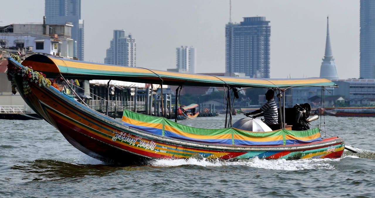 Traditional long-tail boat gliding along the Chao Phraya River in Bangkok, with a striped canopy and city skyline in the background.