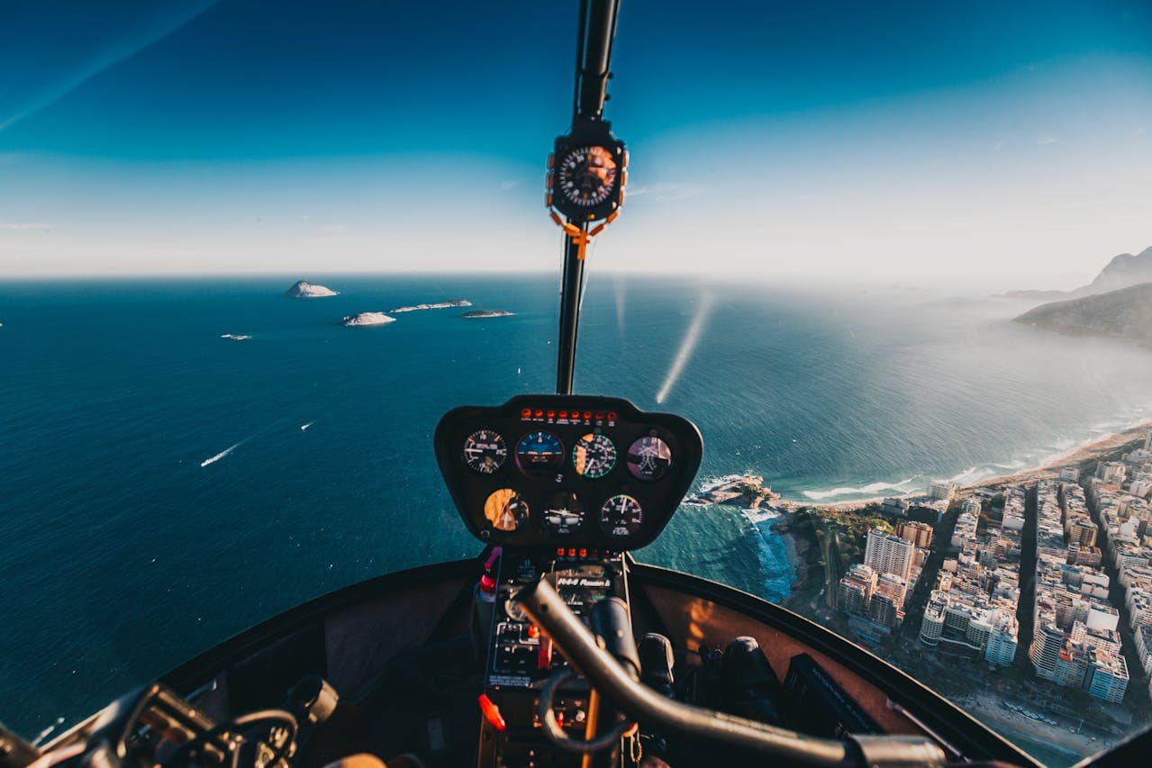 Cockpit view from a helicopter flying over a coastal city with blue ocean, islands, and urban skyline under a clear sky.