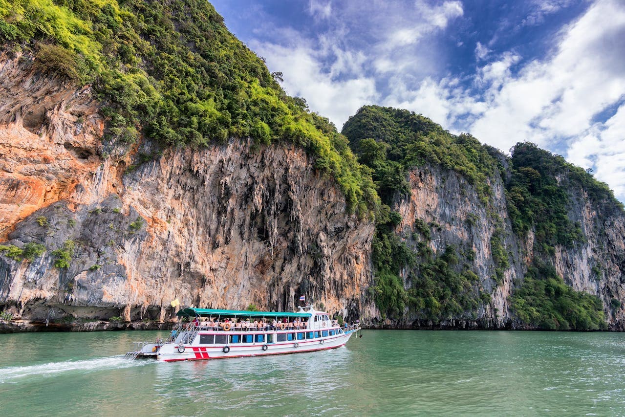 A white and red tour boat with a green canopy cruises on calm green water near towering limestone cliffs covered with lush green vegetation under a partly cloudy blue sky.