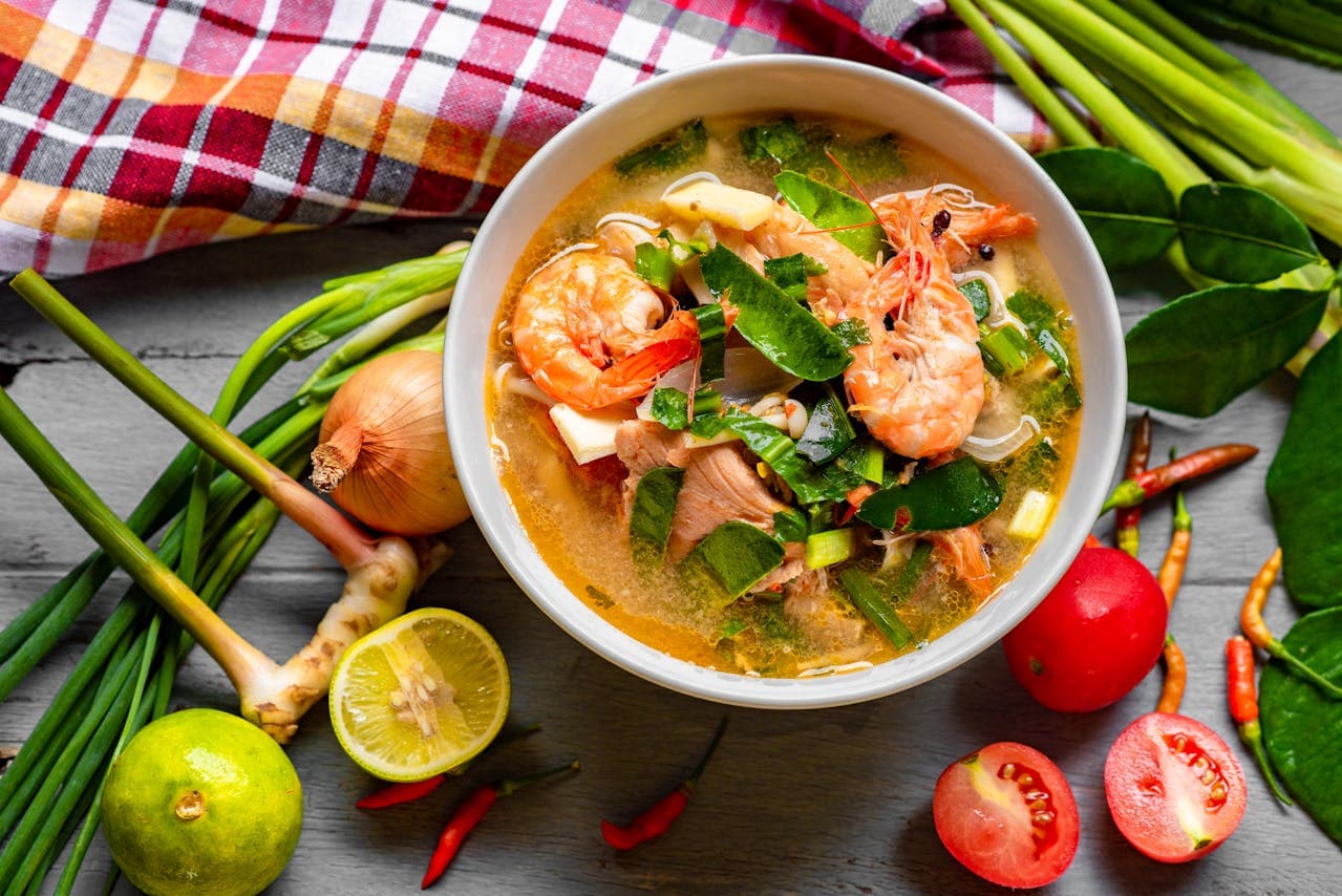 Overhead view of a bowl of Thai Tom Yum soup with prawns, galangal, kaffir lime leaves, spring onions, cherry tomatoes and lime on a rustic wooden table