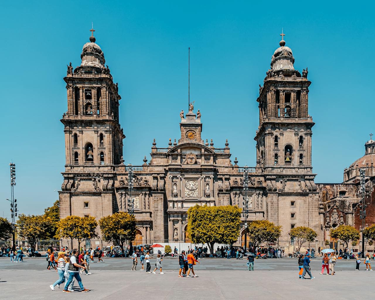 Front façade of the Mexico City Metropolitan Cathedral with its twin bell towers overlooking the Plaza de la Constitución, with visitors strolling under a clear blue sky.