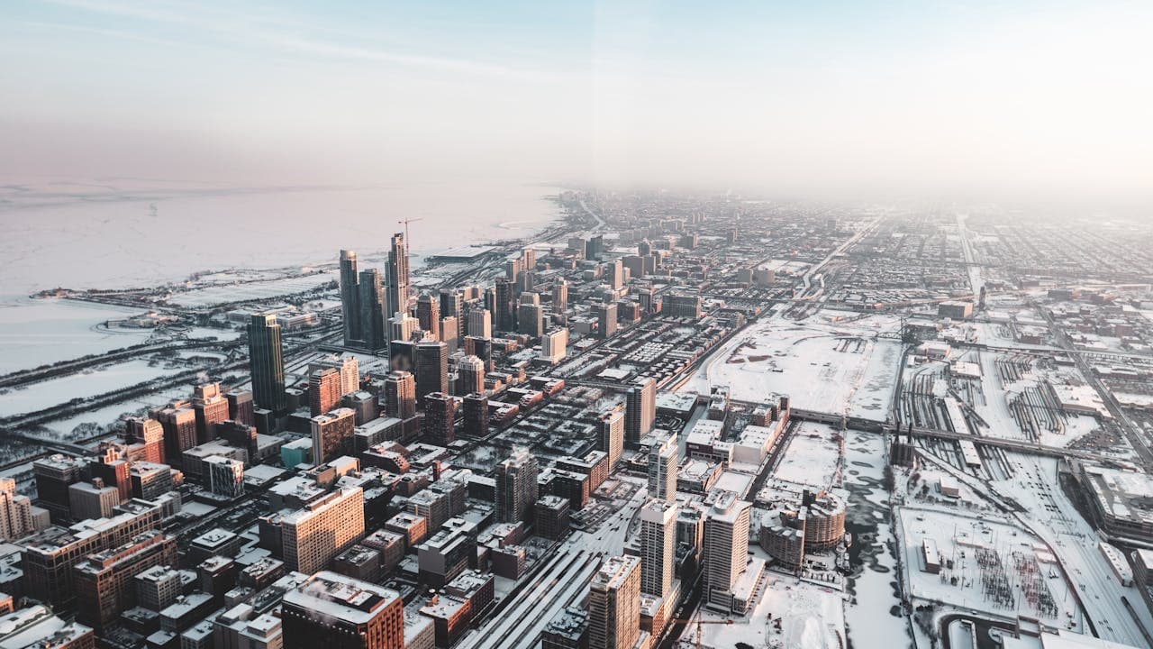 Aerial view of a snow-covered cityscape with skyscrapers, roads, and a distant waterfront under a clear sky.