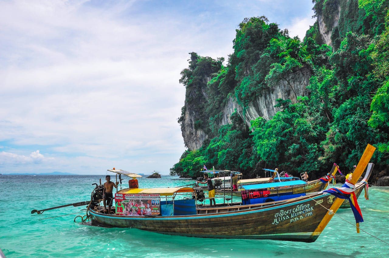 A colorful wooden long-tail boat anchored in crystal-clear turquoise waters against a backdrop of towering limestone cliffs and tropical foliage in southern Thailand.