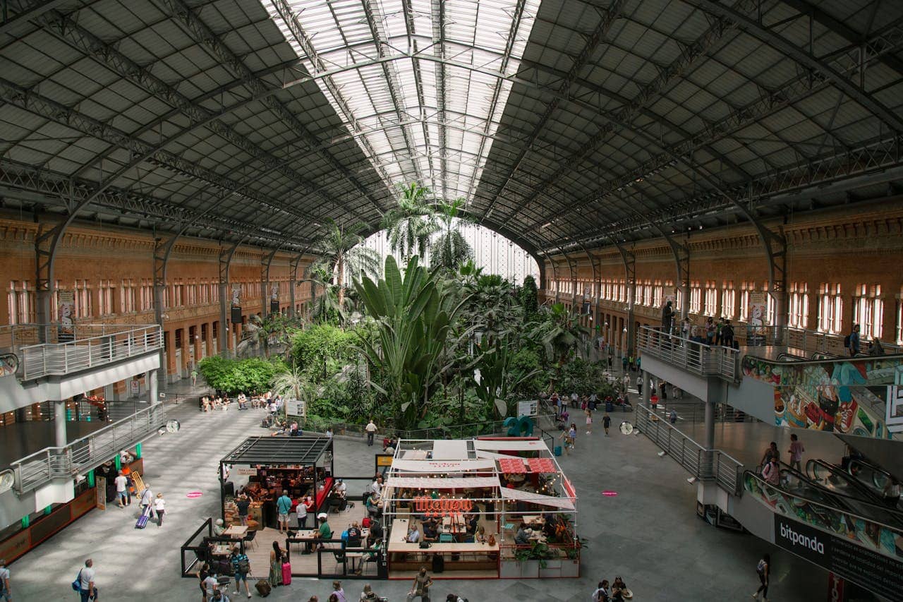 Interior view of Madrid’s Atocha train station featuring a lush indoor tropical garden, high arched glass roof, and people walking through the spacious concourse with shops and cafes.