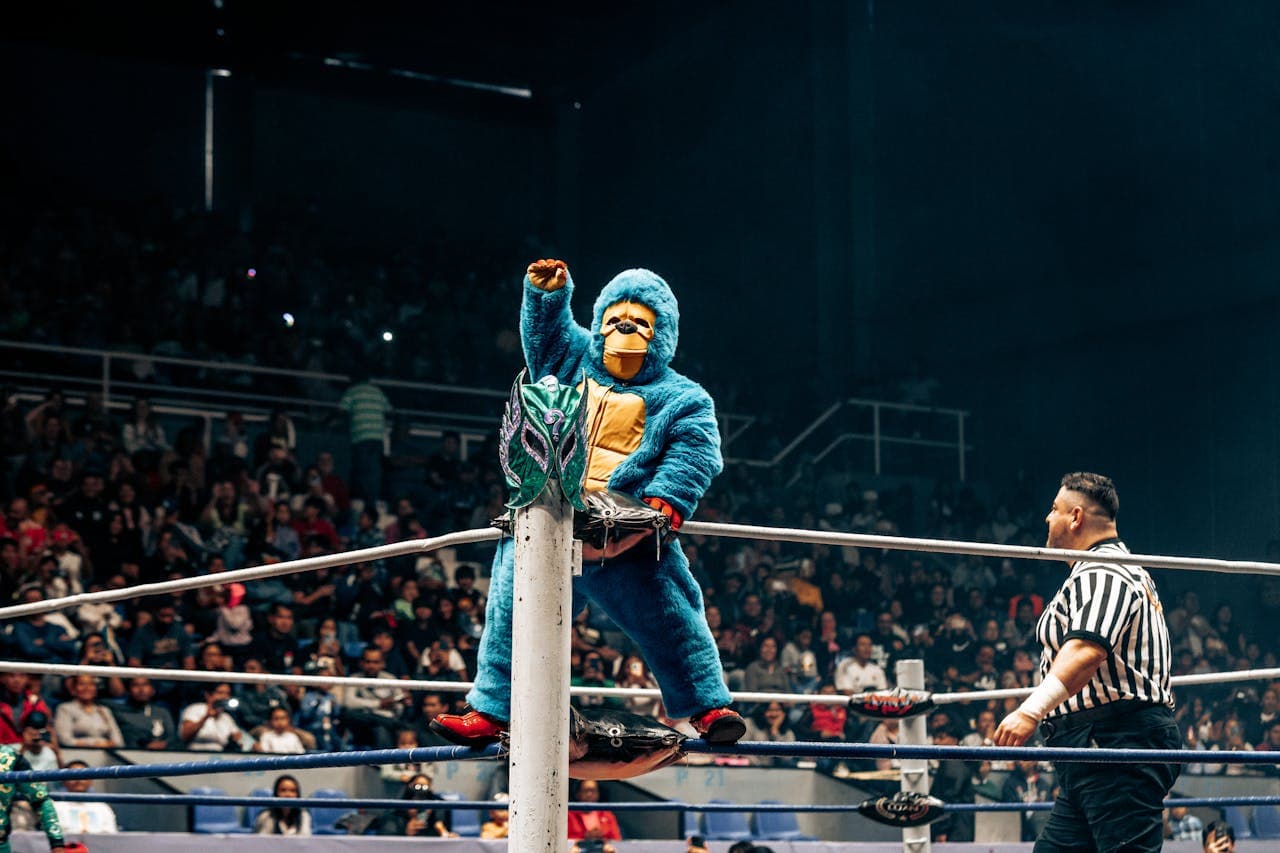 Person in a blue costume standing on the ropes of a wrestling ring in front of a large crowd during a wrestling event.