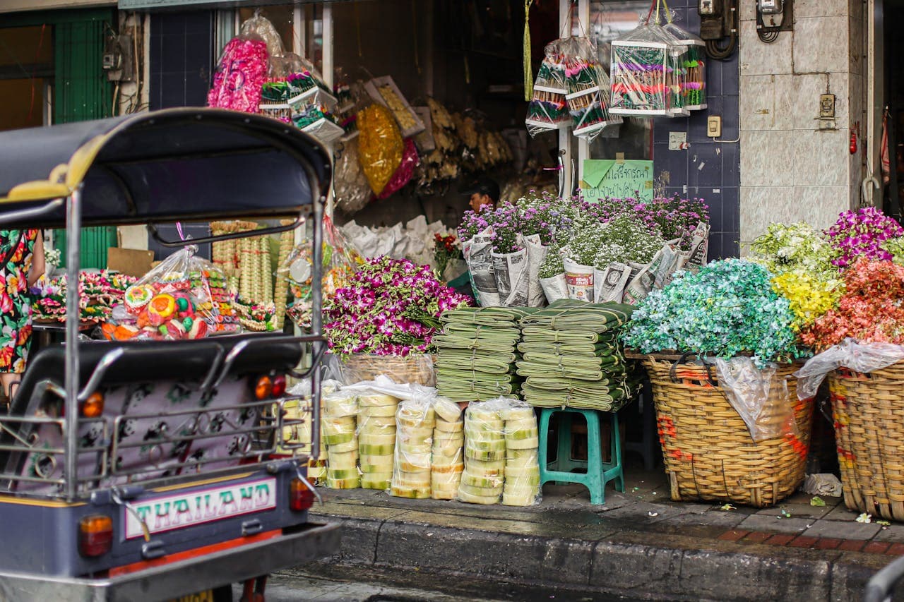 Colorful Thai street market stall with baskets of fresh flowers, folded banana‐leaf offerings wrapped in newspaper, and a parked tuk‐tuk labeled ‘Thailand.’