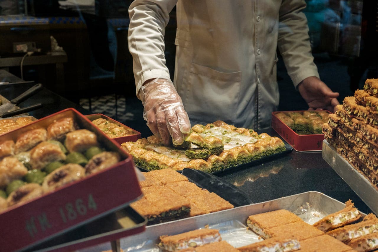 Gloved shop worker arranging trays of baklava and pistachio-filled pastries behind a glass display.