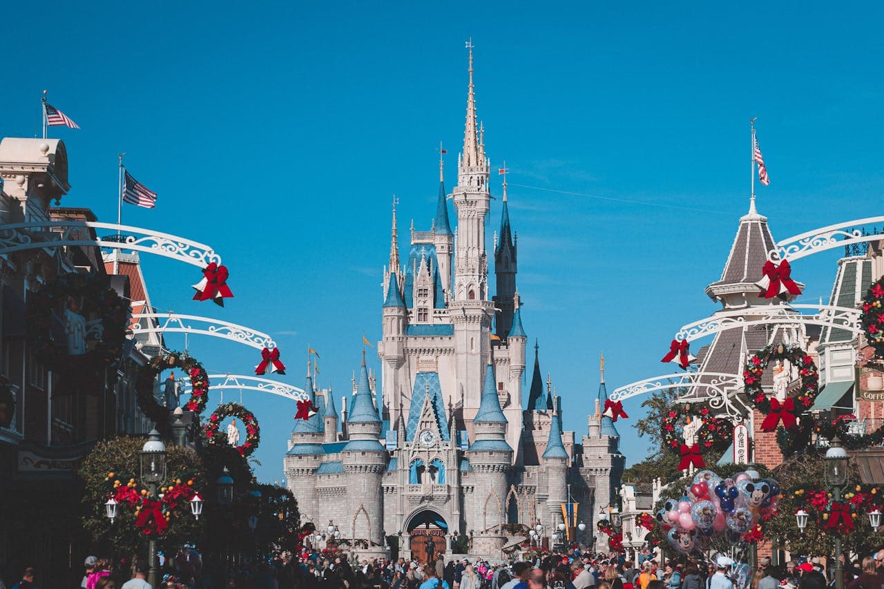 A bustling theme park with a grand castle, festive decorations, and a clear blue sky.