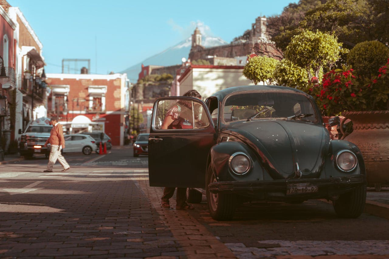 Classic Volkswagen Beetle parked on a cobblestone street in a colorful Mexican town with colonial buildings and a volcano in the background.