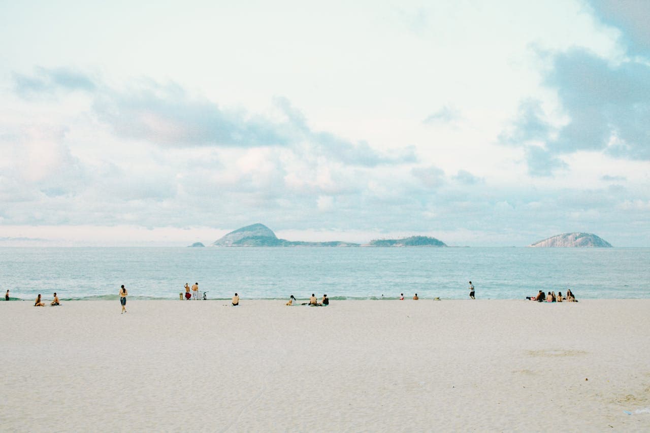 Wide sandy Copacabana Beach in Rio de Janeiro, Brazil, dotted with sunbathers and swimmers and framed by the distant Cagarras Islands under a bright sky.