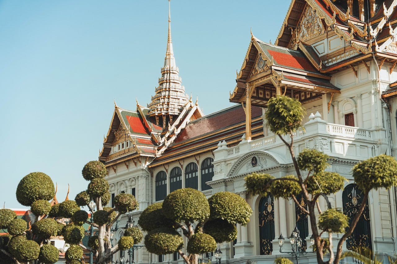 Exterior view of Bangkok’s Grand Palace complex, showcasing ornate gilded spires, red-and-gold tiled roofs, and sculpted topiary in the foreground.