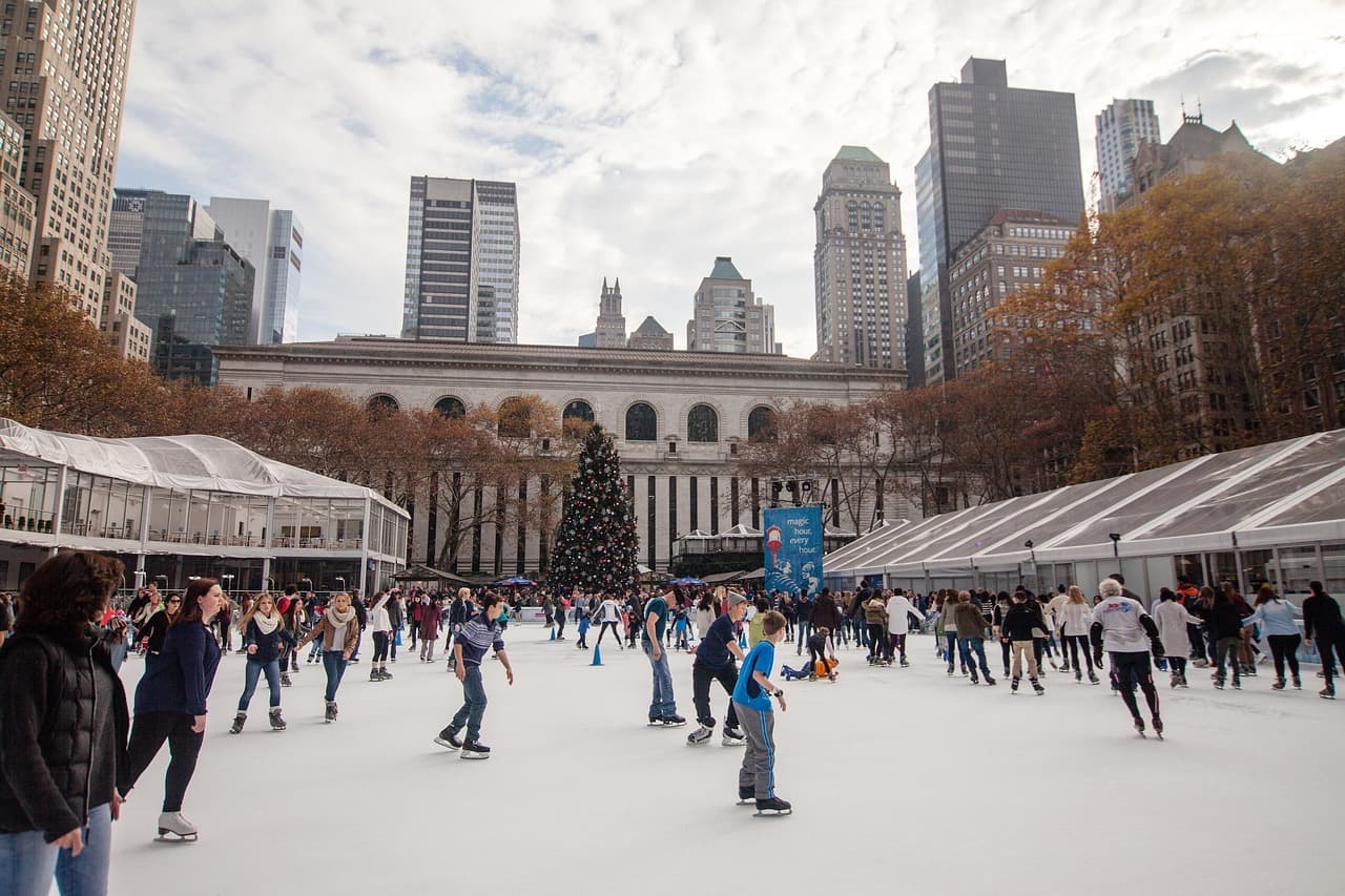 People ice skating at an outdoor rink in a city park, surrounded by tall buildings and a large Christmas tree.