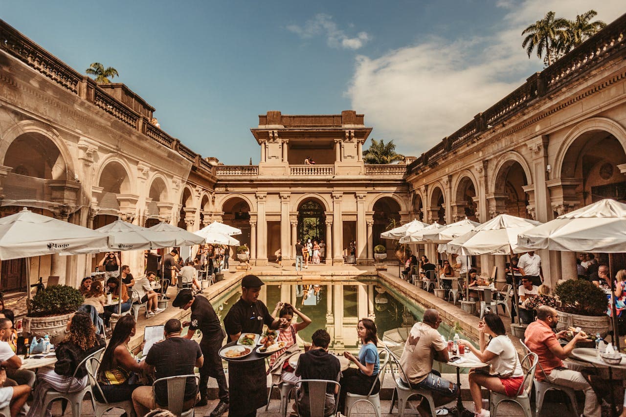 Outdoor café in the sunlit courtyard of Parque Lage, Rio de Janeiro, featuring diners seated around a central reflecting pool beneath white umbrellas, framed by elegant stone archways and tropical greenery.