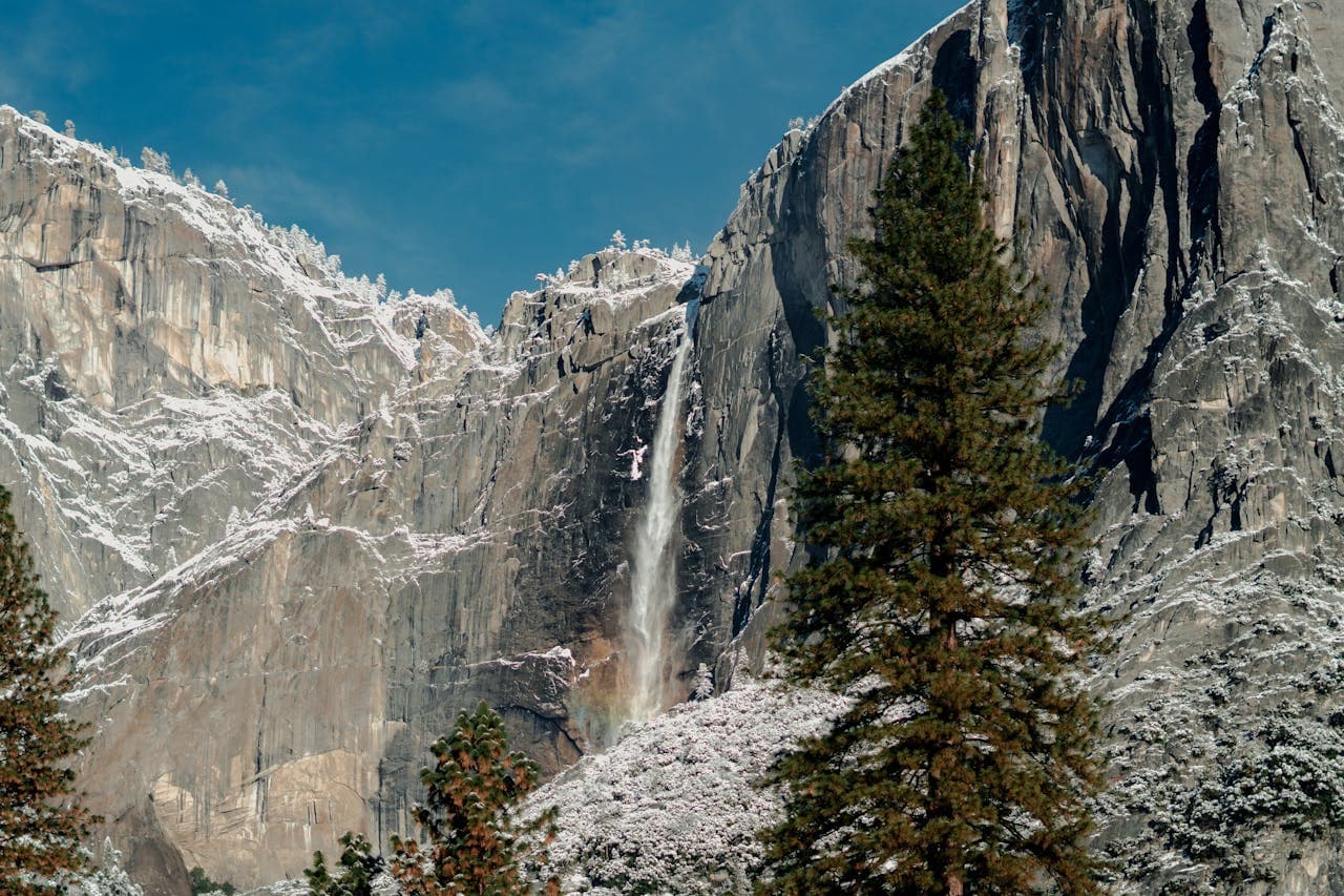 Snow-dusted cliffs with a waterfall cascading down, surrounded by evergreen trees under a clear blue sky.