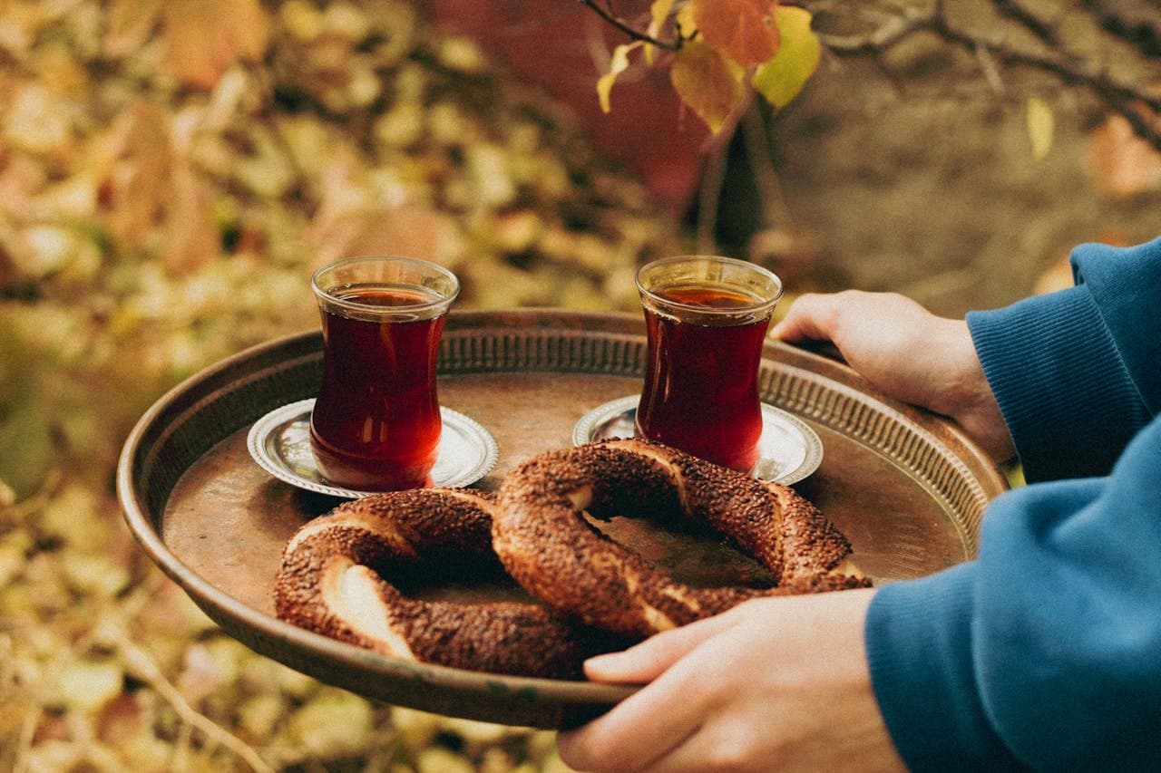 Person's hands in blue sleeves holding a tray with two Turkish tea glasses and sesame simits among autumn leaves.