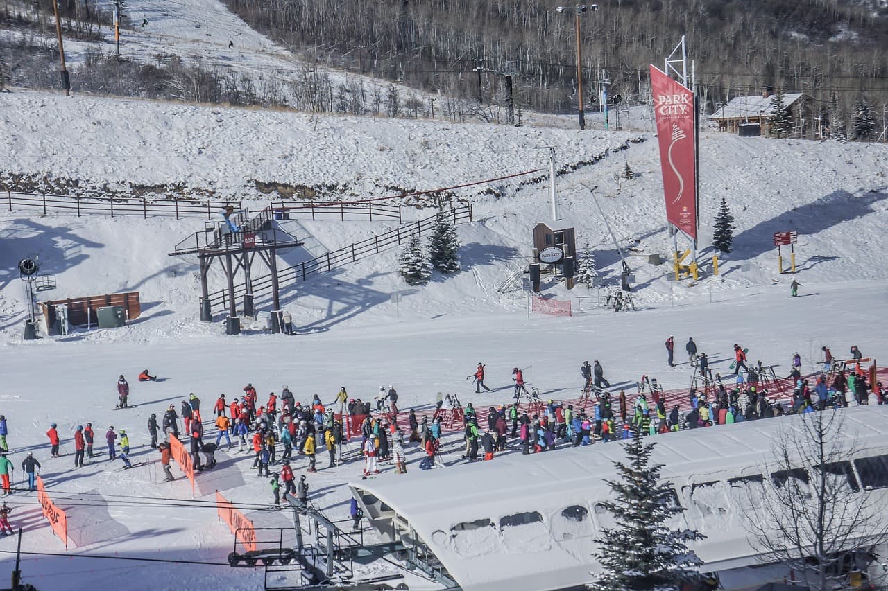 A bustling ski resort with numerous skiers and snowboarders on snowy slopes, surrounded by trees and buildings under a clear blue sky.