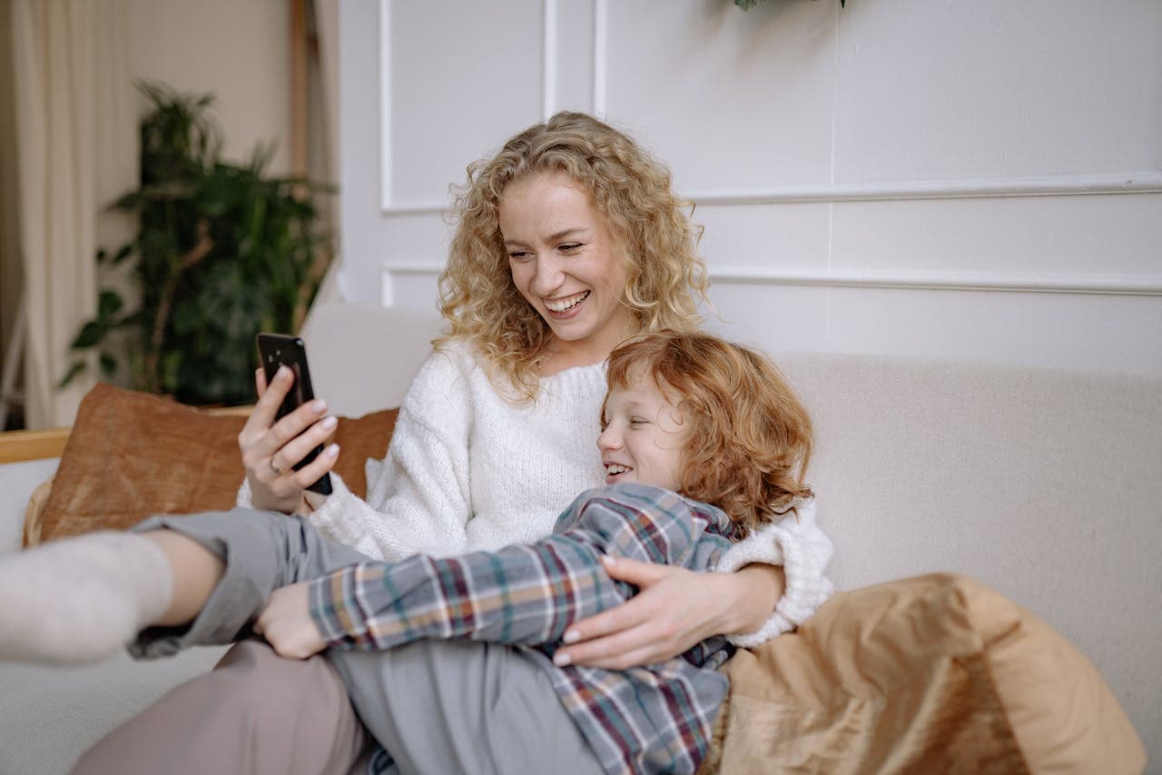 Smiling woman with curly hair sits on a couch, holding a phone, while a laughing child with red hair lies on her lap.