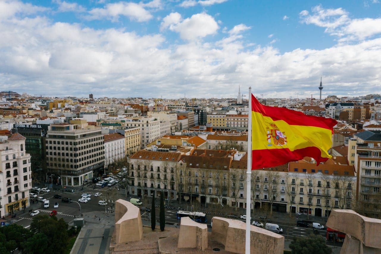 Panoramic view of Madrid city skyline with the Spanish flag waving prominently in the foreground under a partly cloudy sky.