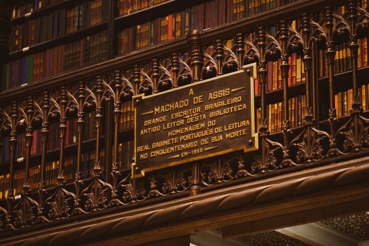 Ornate iron-work balcony and shelves of antique books inside the Royal Portuguese Cabinet of Reading library in Rio de Janeiro, Brazil, with a dedication plaque to Machado de Assis.