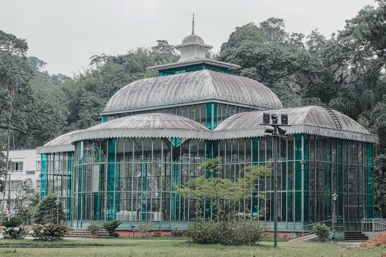 Wrought-iron and glass greenhouse in the Rio de Janeiro Botanical Garden, with three domed roofs and intricate Victorian-era latticework, set amidst lush Atlantic Forest vegetation.