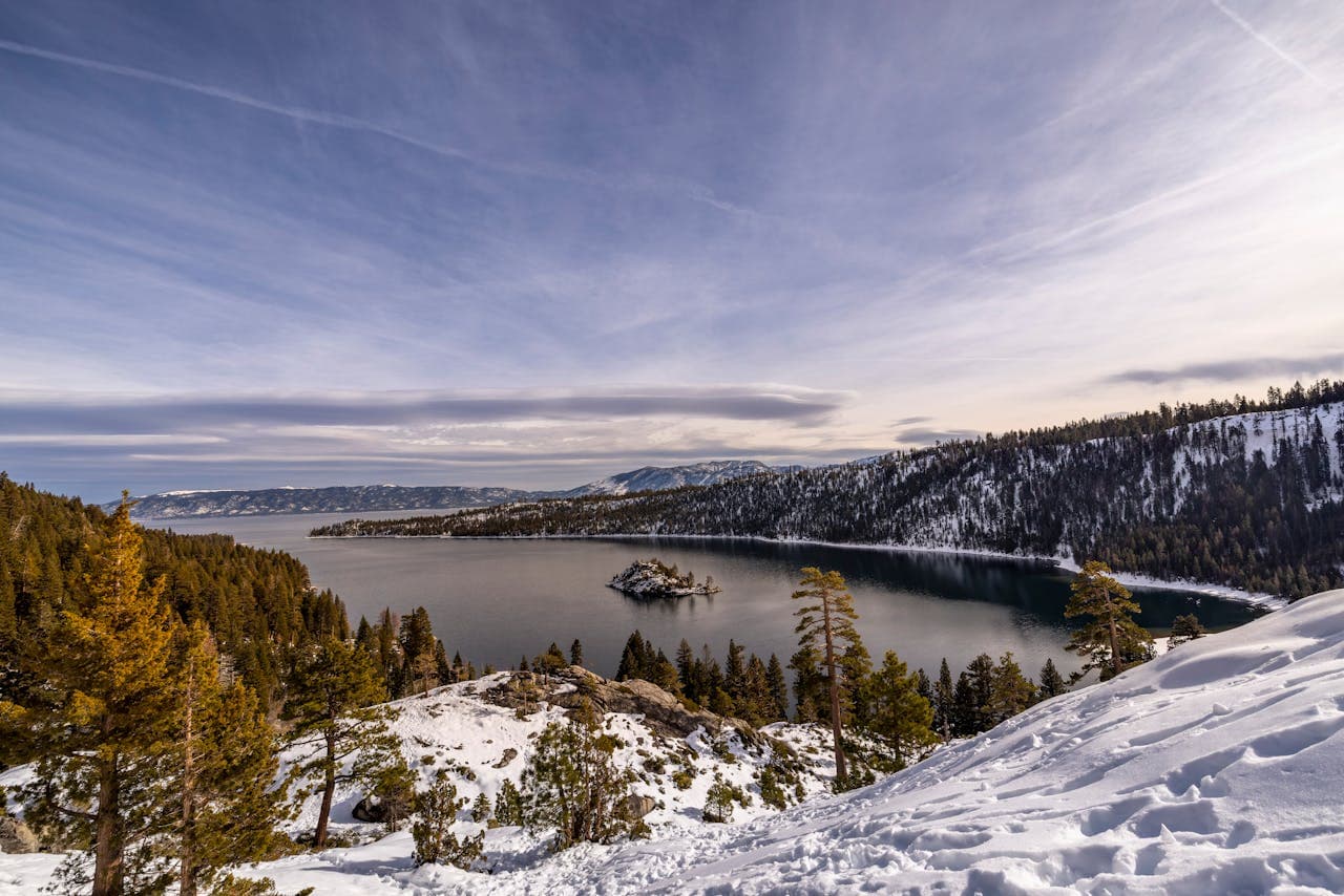 Snowy landscape overlooking a lake surrounded by pine trees and mountains under a partly cloudy sky.
