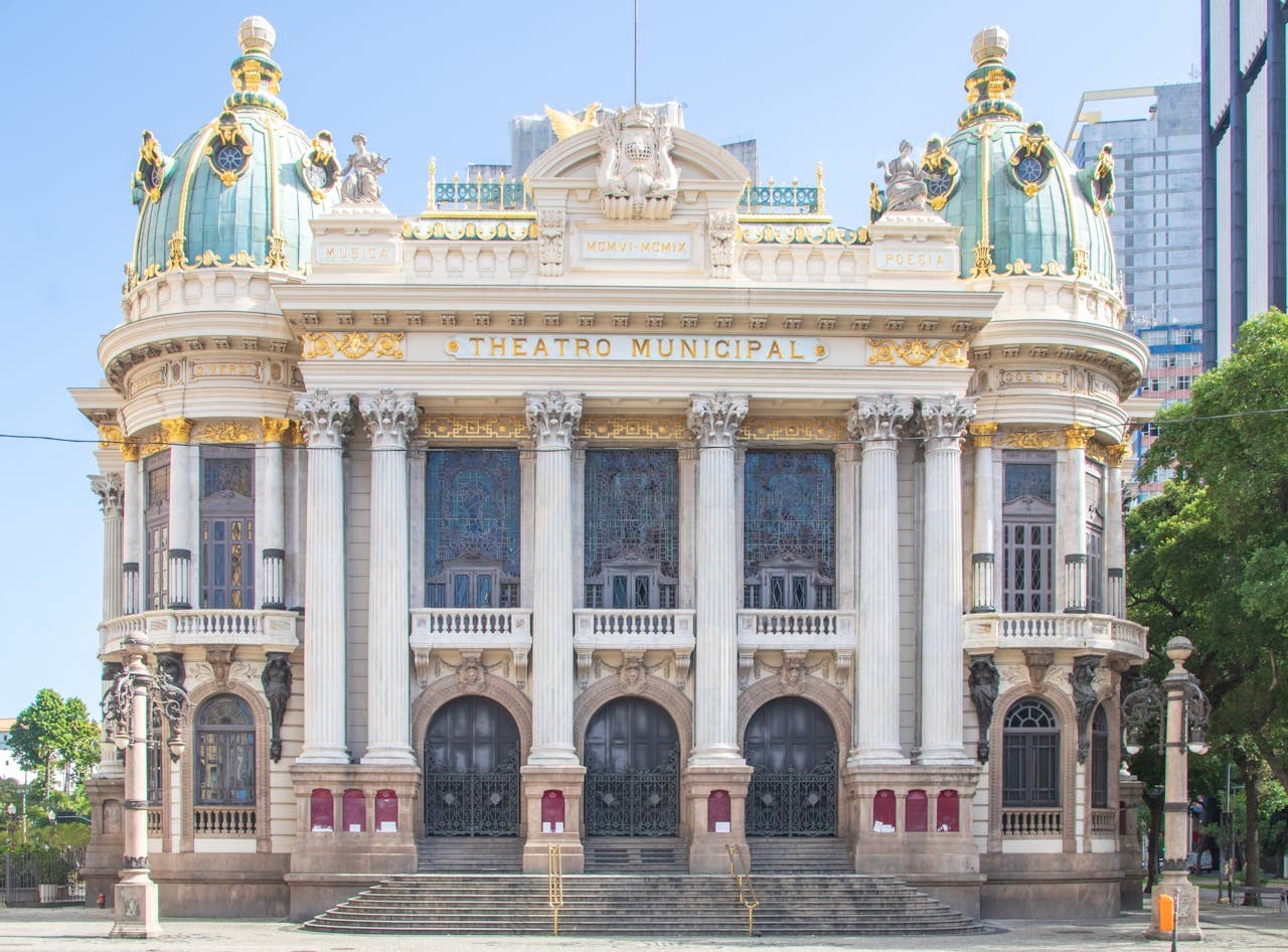 Front façade of the historic Theatro Municipal in Rio de Janeiro, showcasing its ornate green-and-gold domes, Corinthian columns, intricate sculptures, and gilded Neoclassical detailing against a clear blue sky.