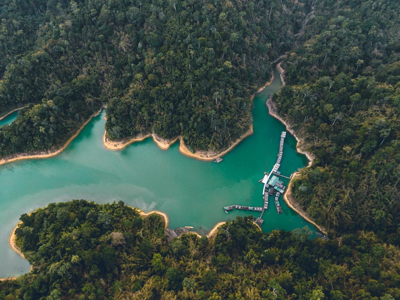 A winding emerald-green reservoir snaking through dense jungle, with a cluster of floating bungalows and docks nestled along the forested shoreline.