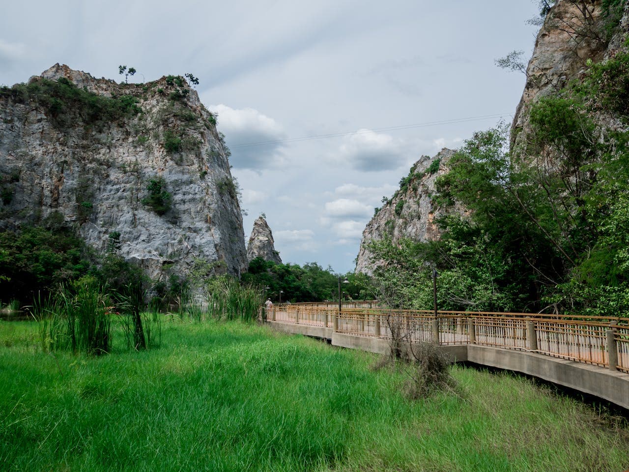 Scenic view of Khao Ngu Stone Park with lush green grass, rocky limestone mountains, and a wooden walkway in Ratchaburi, Thailand.