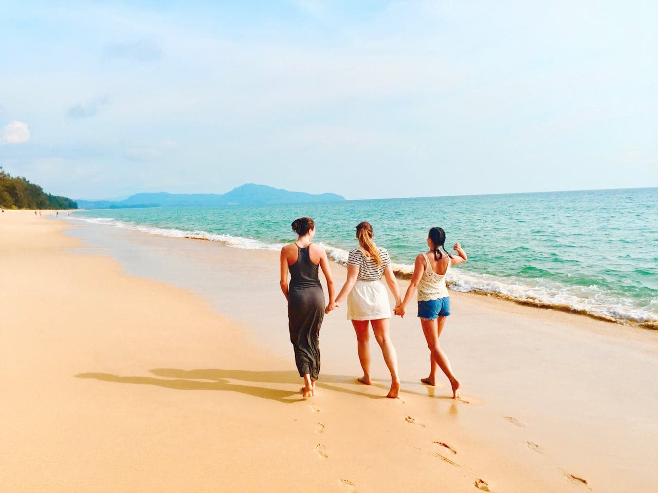 Three women walking hand in hand along a sandy beach with clear blue water and mountains in the background on a sunny day.
